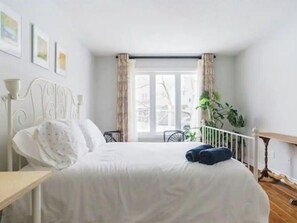 Cozy retreat with a neatly made bed, bedside table, and comfortable chair, featuring a large window with curtains and a potted plant.