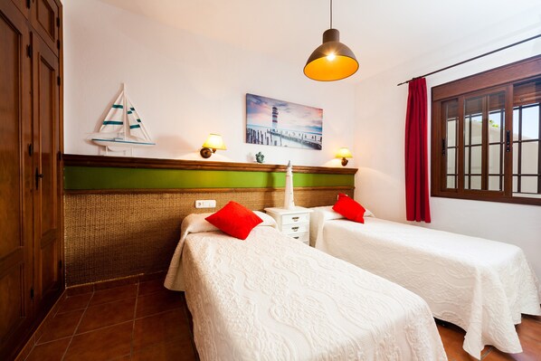 Habitación de hotel con dos camas, una lámpara de pared, decoración con un velero, un cuadro de un faro y una ventana con cortinas rojas.