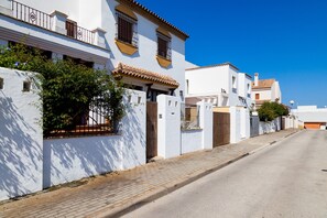 Une rue bordée de bâtiments blancs aux toits de tuiles et aux balcons, avec un trottoir pavé et un ciel d’un bleu limpide.