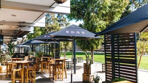 An outdoor patio area with wooden tables and chairs, covered by umbrellas, and surrounded by greenery. - 🔆PINEPLACE🔆BFast,Wi-Fi,Walk2Pizza,Coffee,Liquor (Norlane)