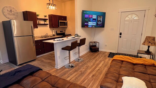 A modern living room with a brown leather sofa, a small kitchen area with a refrigerator and microwave, a dining table with two bar stools, and a TV mounted on the wall.