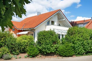 A two-story house with a red-tiled roof, surrounded by lush greenery.