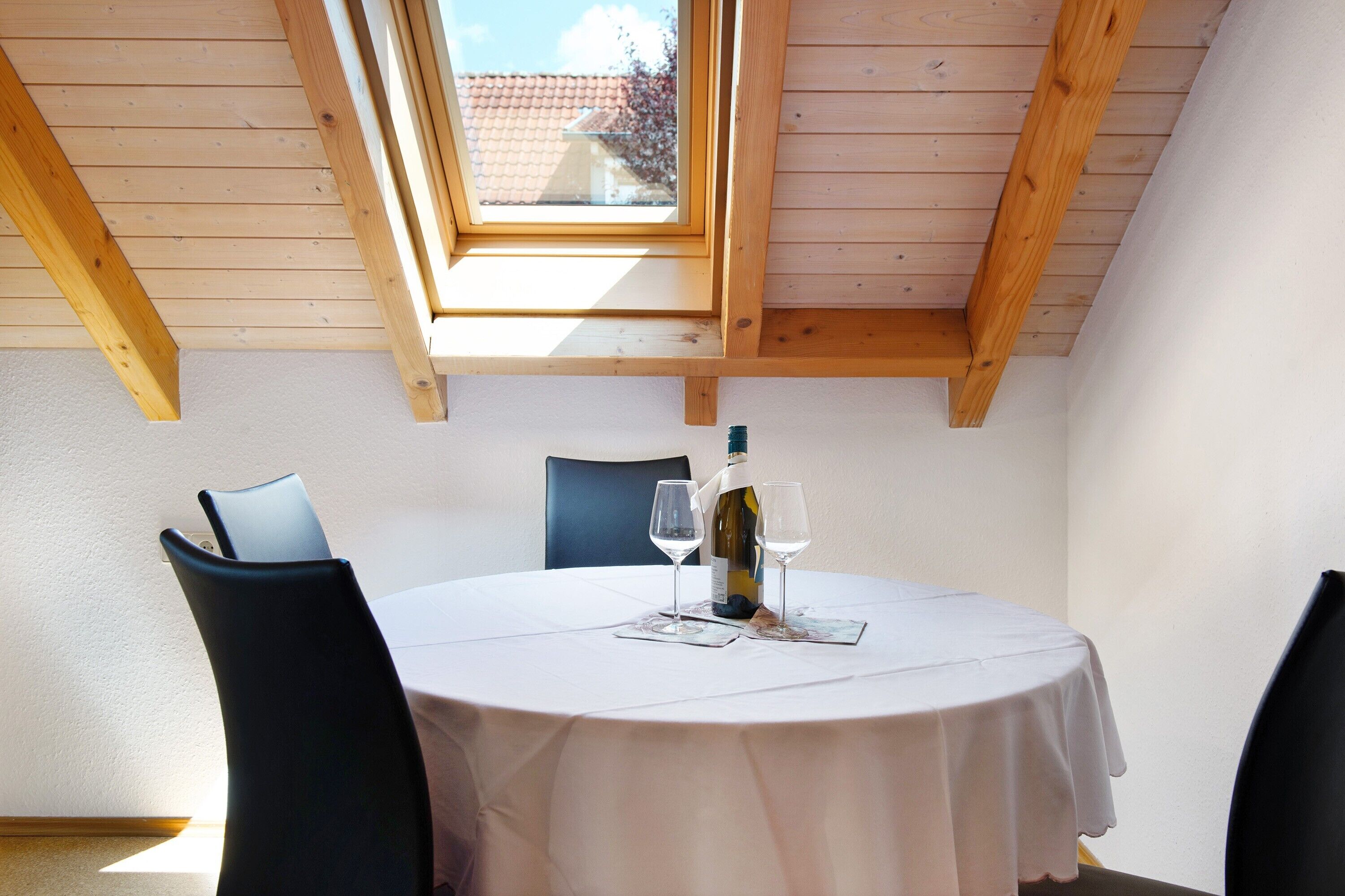 A round table with a white tablecloth, two wine glasses, and a bottle of wine, set in a room with wooden beams and a skylight.