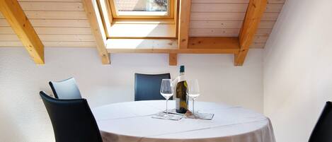 A round table with a white tablecloth, two wine glasses, and a bottle of wine, set in a room with wooden beams and a skylight.