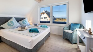 A bedroom with a bed, a wicker chair, a desk, and a window with a view of a house. - Holiday Home 'Ankerplatz' with Sea View, Sauna, and Gas Fireplace (Dranske)