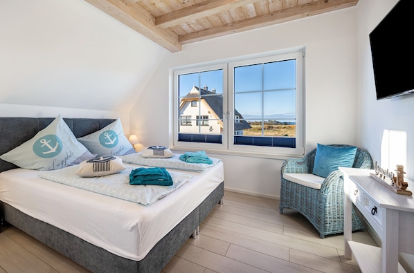 A bedroom with a bed, a wicker chair, a desk, and a window with a view of a house.