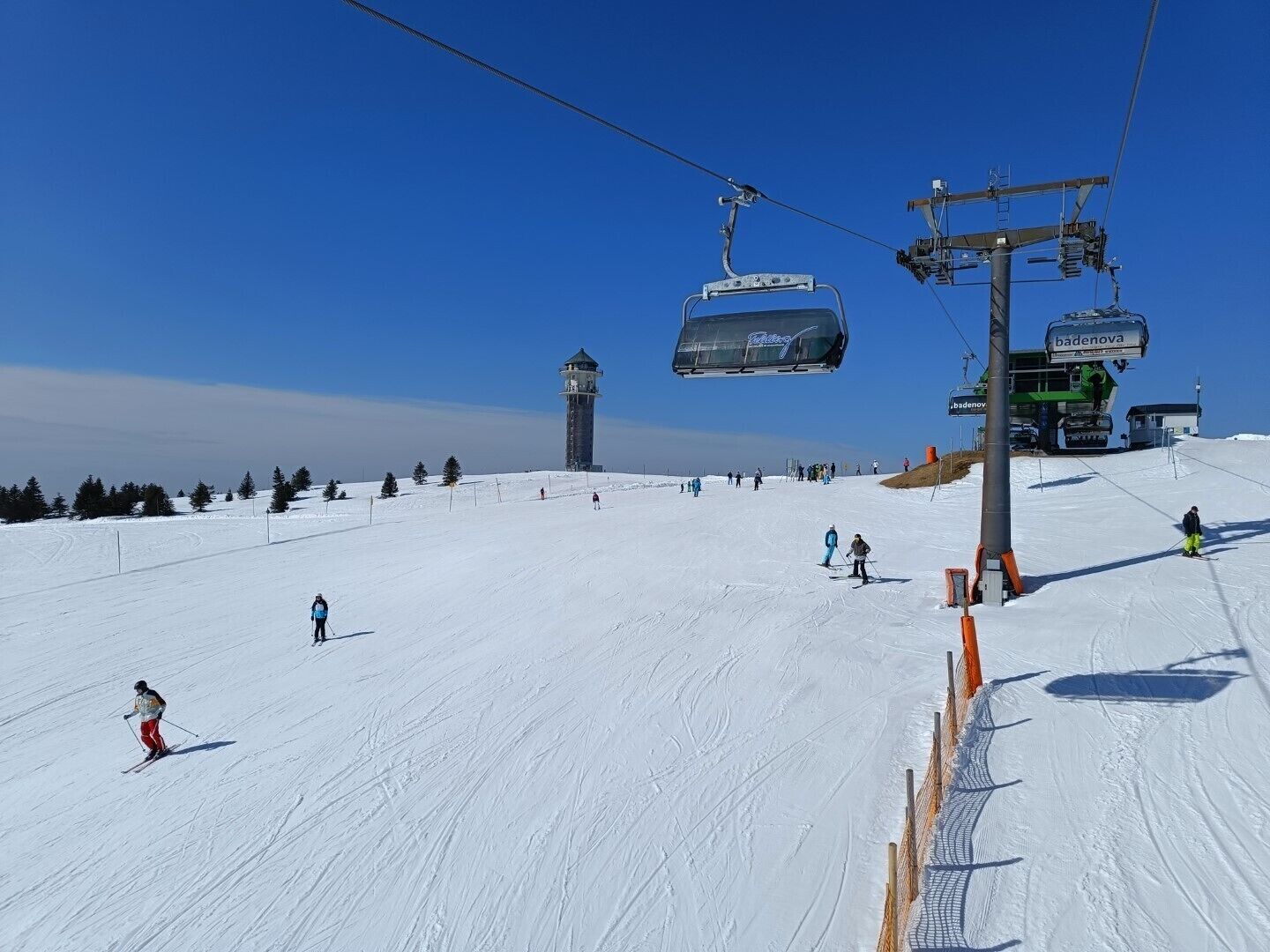 Eine Skipiste mit Skifahrern, einer Sesselbahn und einem Turm im Hintergrund.