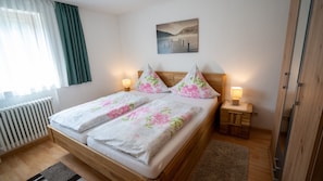 A double bed with floral bedding, two bedside tables with lamps, a radiator, and a painting on the wall. - Holiday Apartment "Haus Franziska" with Mountain View, Private Terrace & Wi-Fi (Todtnau /Brandenberg)