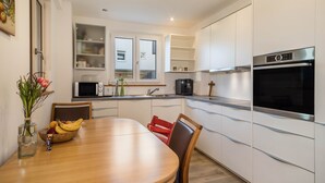 Una cocina moderna con armarios blancos, horno empotrado y comedor con mesa y sillas de madera.