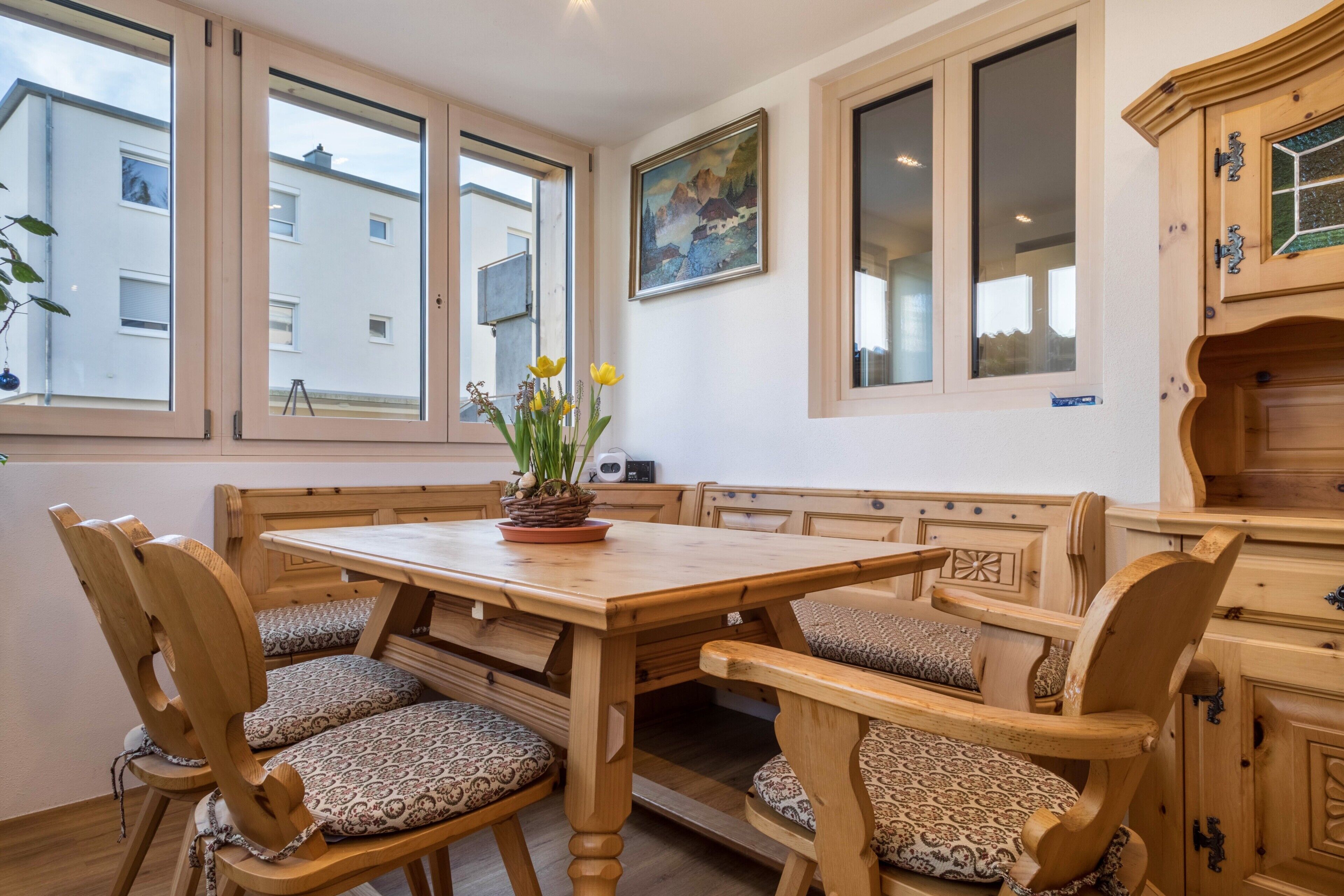 Un espace repas comprenant une table et des chaises en bois, un banc et un pot de fleurs avec des fleurs jaunes.