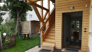 Una escalera de madera que conduce a una puerta, un pequeño espacio al aire libre con una mesa y sillas, y un jardín con una planta en maceta.