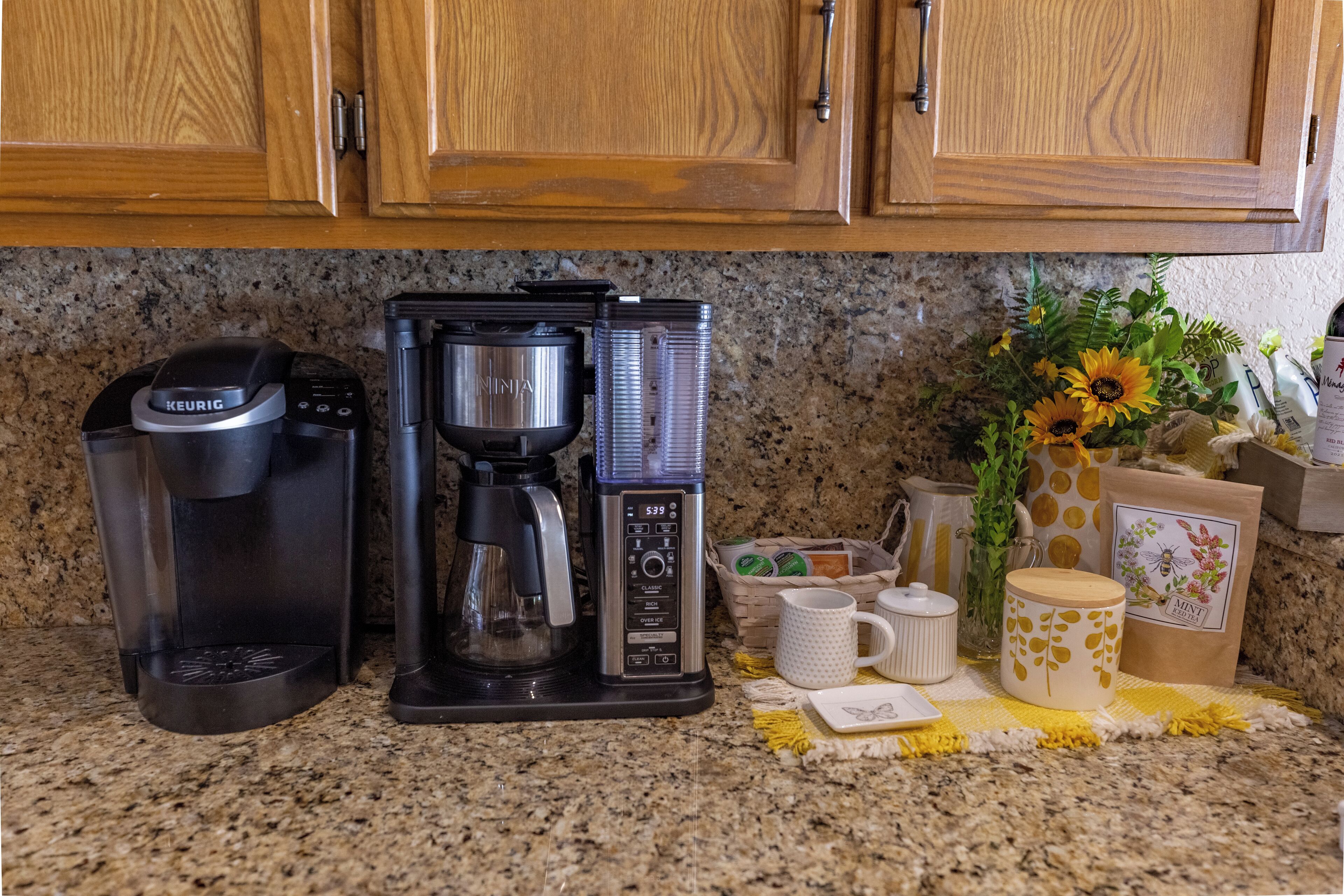 Une cuisine équipée d’une machine à café, d’un distributeur d’eau et d’une cafetière. On y trouve des tasses, un vase avec des fleurs et un sac de grains de café sur le comptoir.