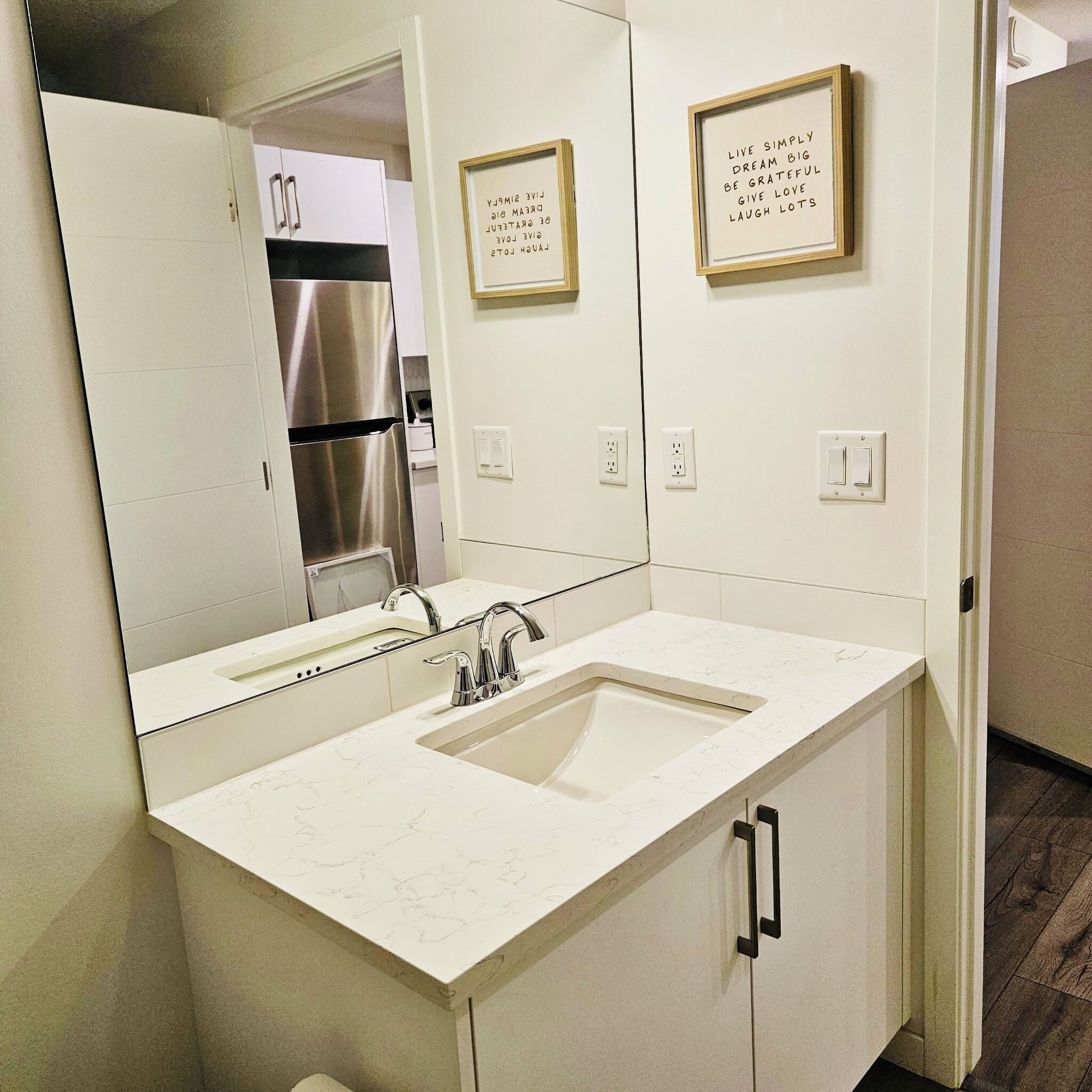 A modern bathroom with a white sink, stainless steel refrigerator in the background, and motivational quotes on the wall.