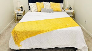 Un lit bien fait, recouvert d’une couvre-lit blanche et jaune, avec une tête de lit bleu foncé et plusieurs oreillers aux motifs variés. On y trouve deux tables de chevet, chacune avec une lampe, et une horloge sur l’une d’elles. La pièce est dotée d’un sol recouvert d’une moquette et de murs aux couleurs neutres.