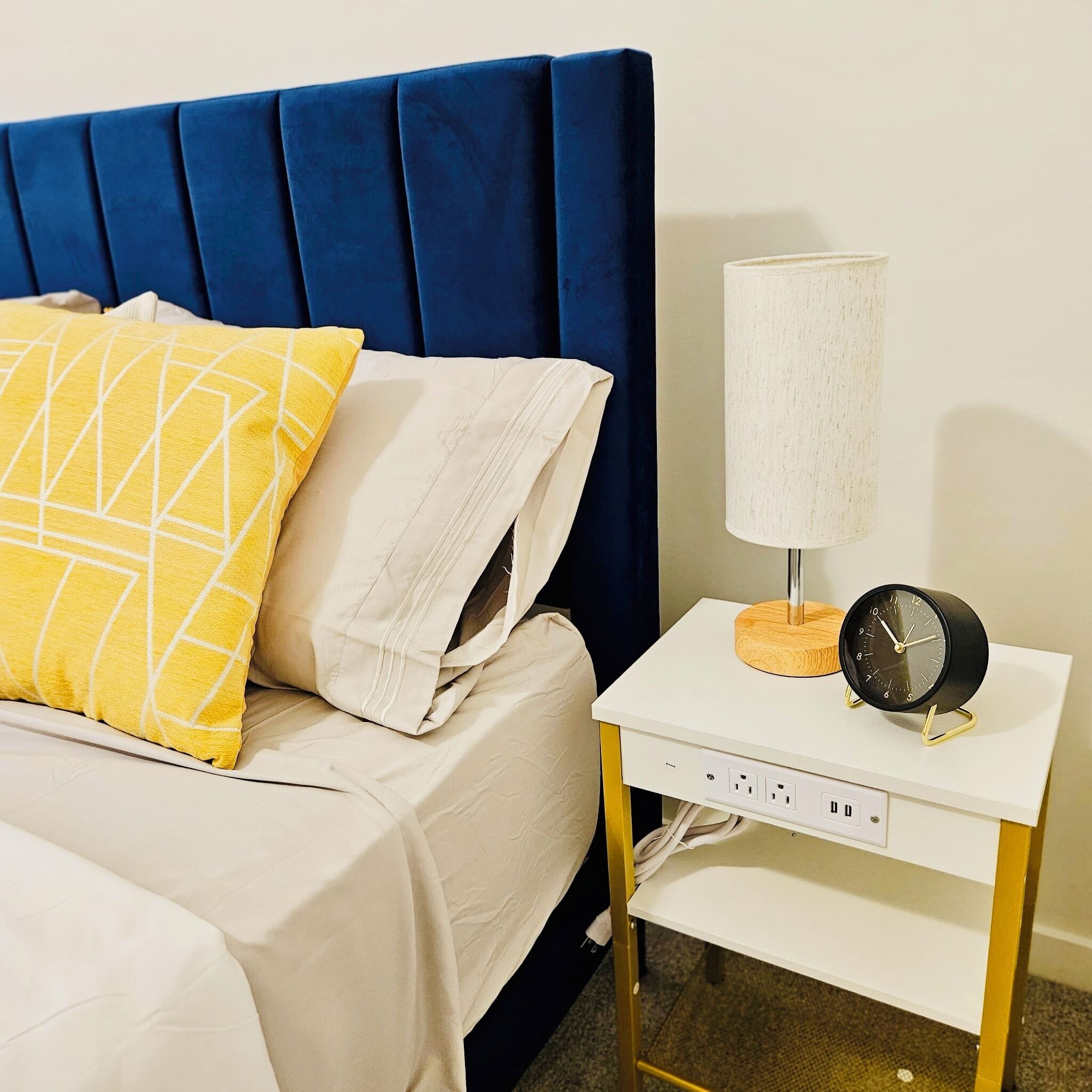 A neatly made bed with a blue headboard, a yellow pillow, and a white pillow. There is a bedside table with a lamp and a clock.