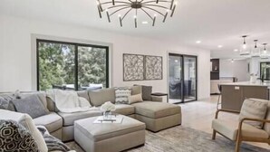 A modern living room with a sectional sofa, ottoman, and a dining area with a kitchen in the background.