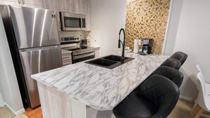 A modern kitchen with a stainless steel refrigerator, microwave, and oven. There is a marble countertop with a sink and black bar stools.
