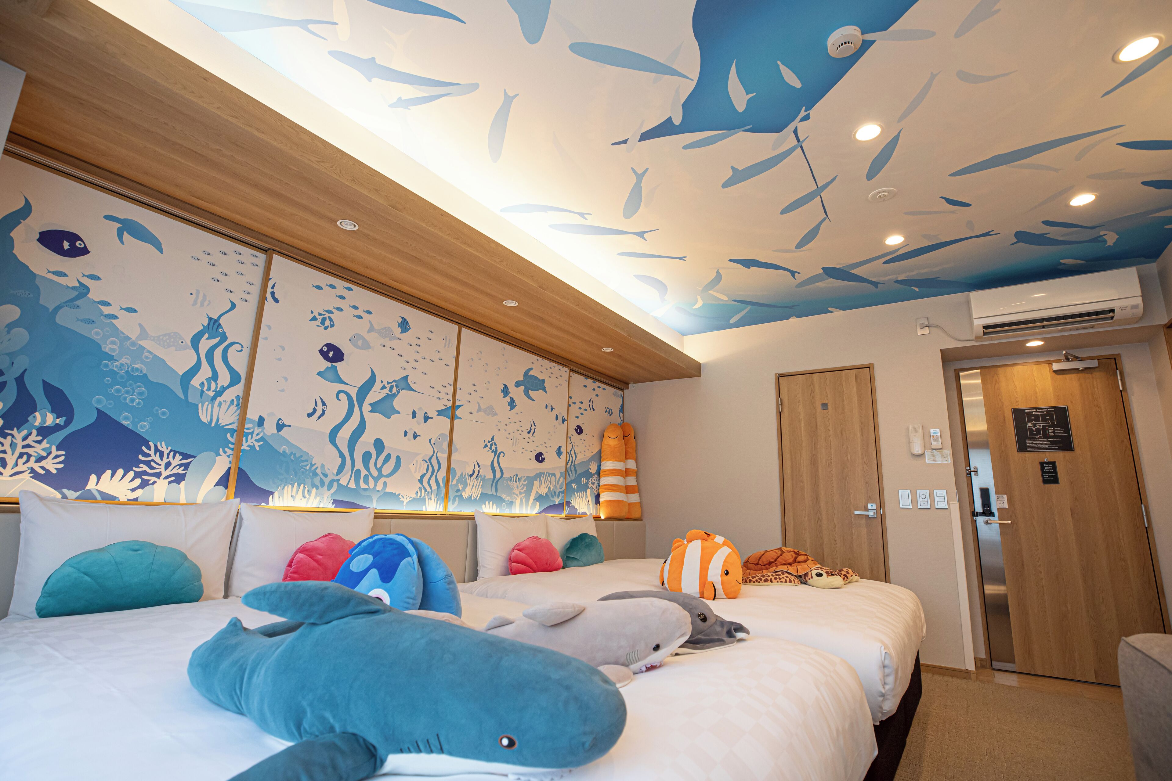 Habitación de hotel con un mural en el techo de peces y vida marina, una cama con almohadas de tiburón y delfín, y un cabecero de madera.