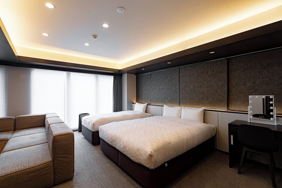 A modern hotel room with two beds, a sofa, a desk with a mirror, and a large window with blinds.