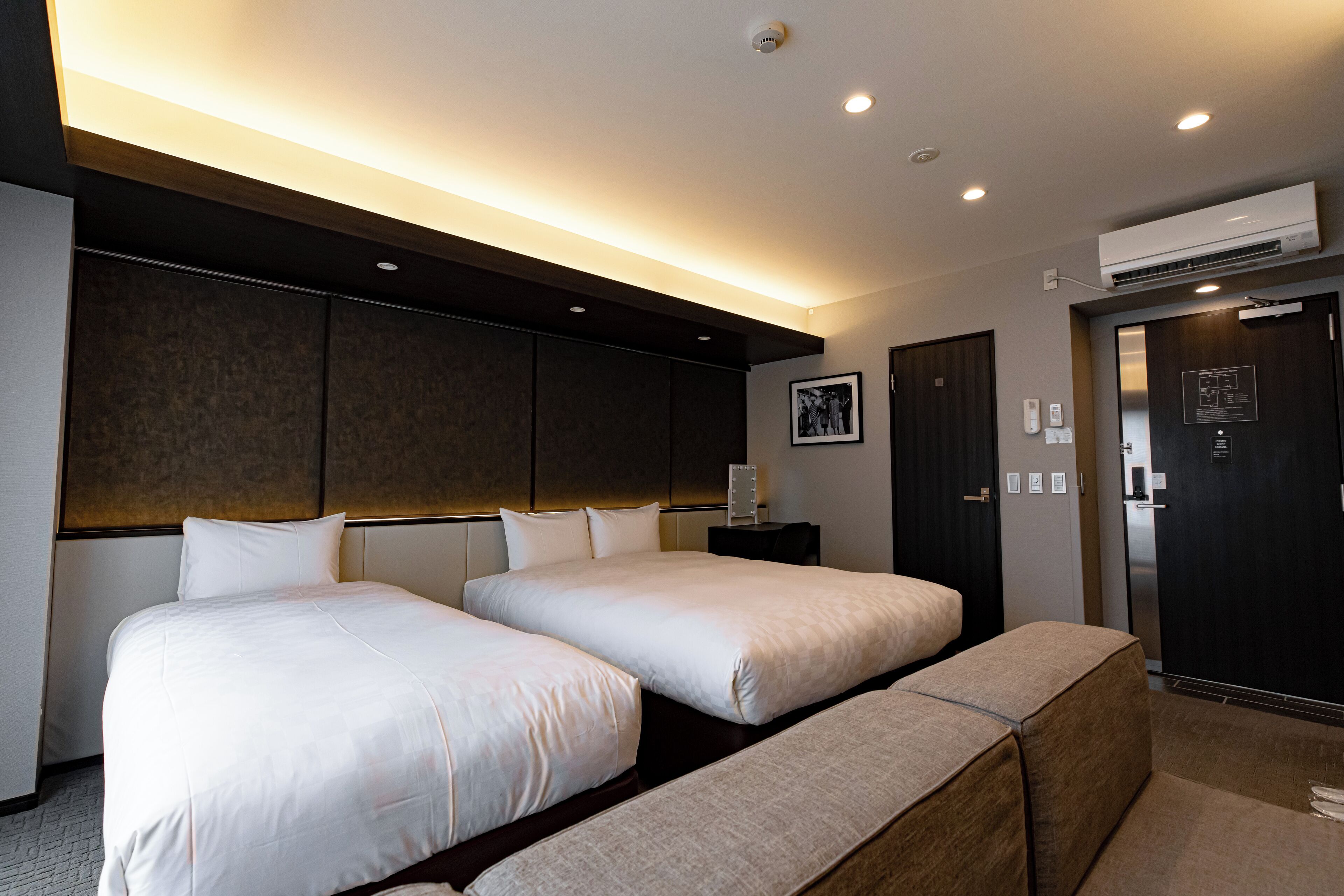 A hotel room with two beds, a bench, a bedside table, an air conditioning unit, and a door with a lock.