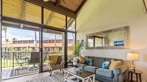 A spacious living area with a large sliding glass door leading to a balcony, a grey sofa, a coffee table, and a small side table with a lamp. - Perch - Luana Kai Loft: Captivating Modern 2-Bedroom for a Dream Vacation (Kihei)