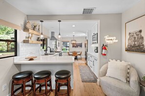 A modern kitchen with a central island, bar stools, a dining area, and a cozy seating area with a fire extinguisher on the wall.