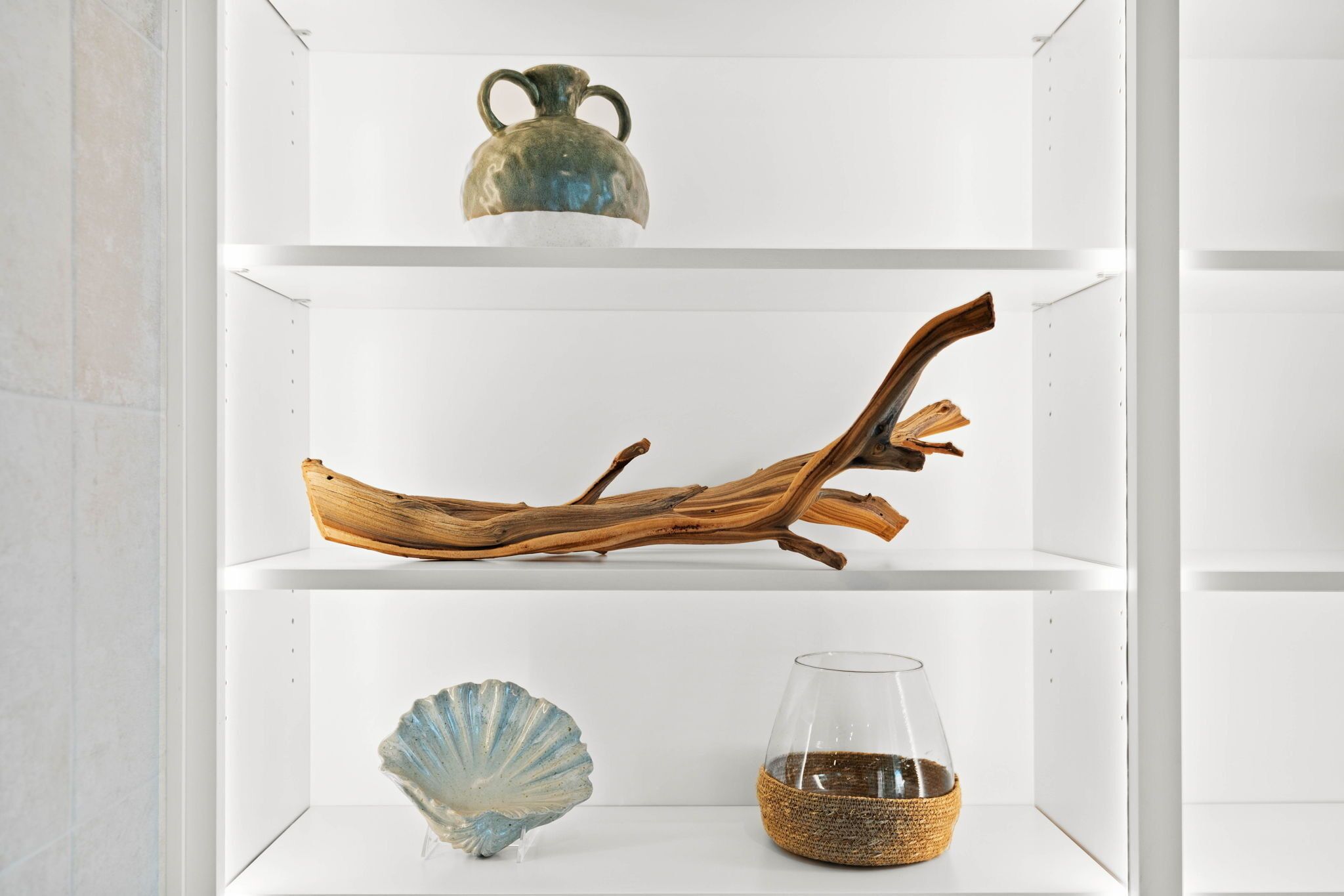 A white shelving unit with three shelves. The top shelf holds a ceramic vase, the middle shelf displays a piece of driftwood, and the bottom shelf features a glass vase with a woven base.