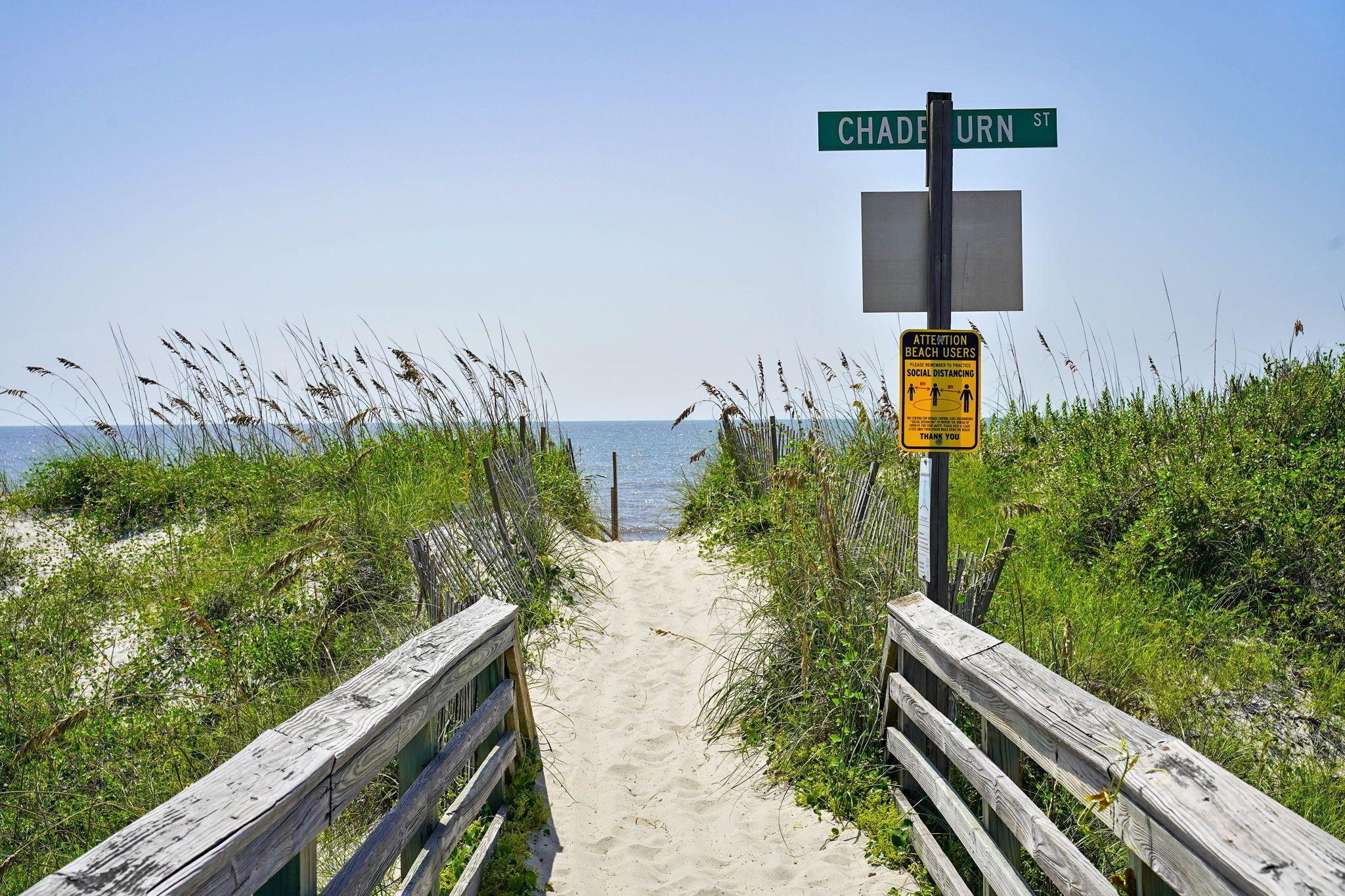 Un sentier bordant la plage, agrémenté d’une clôture en bois, un panneau de signalisation indiquant la rue Chadburn et un panneau rappelant les mesures de distanciation sociale.