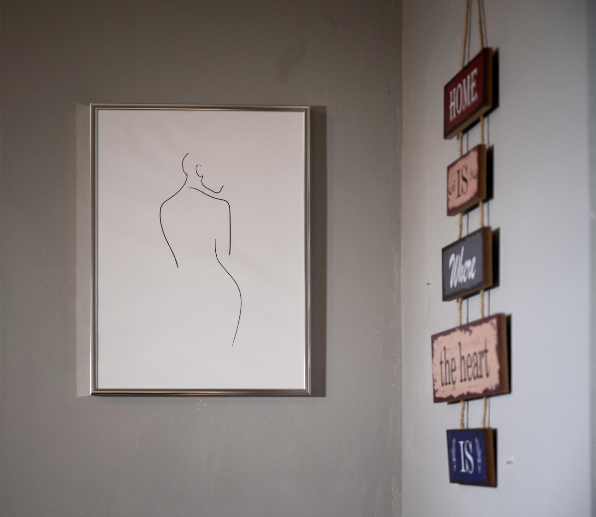 A framed line drawing of a human figure on a wall, next to a wall with hanging signs reading "HOME," "IS," "Where," "the heart," and "IS."