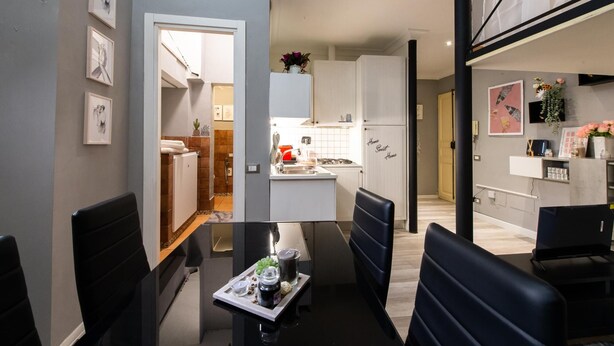 Una cocina moderna con armarios blancos, una zona de comedor con mesa y sillas negras, y una sala de estar con sofá y televisor.