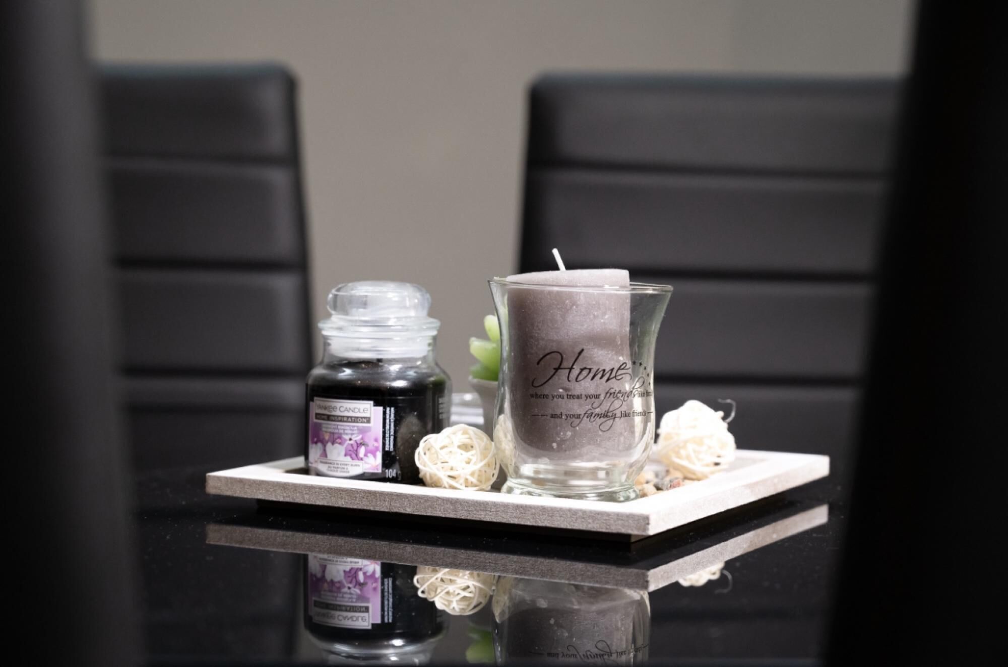 A candle in a glass holder with a "Home" inscription, a small bottle, and decorative items on a reflective surface.