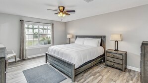 Un dormitorio con cama, mesita de noche, ventilador de techo, ventana con cortinas y suelo de madera.