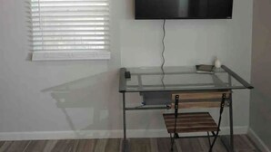 Une pièce équipée d’un téléviseur fixé au mur, d’un bureau en verre, d’un bureau réglable en hauteur et d’une fenêtre avec des stores.