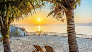 A beach with two wooden lounge chairs, palm trees, and a sunset over the water. - Inlight Lombok Eco Resort (Kuta)