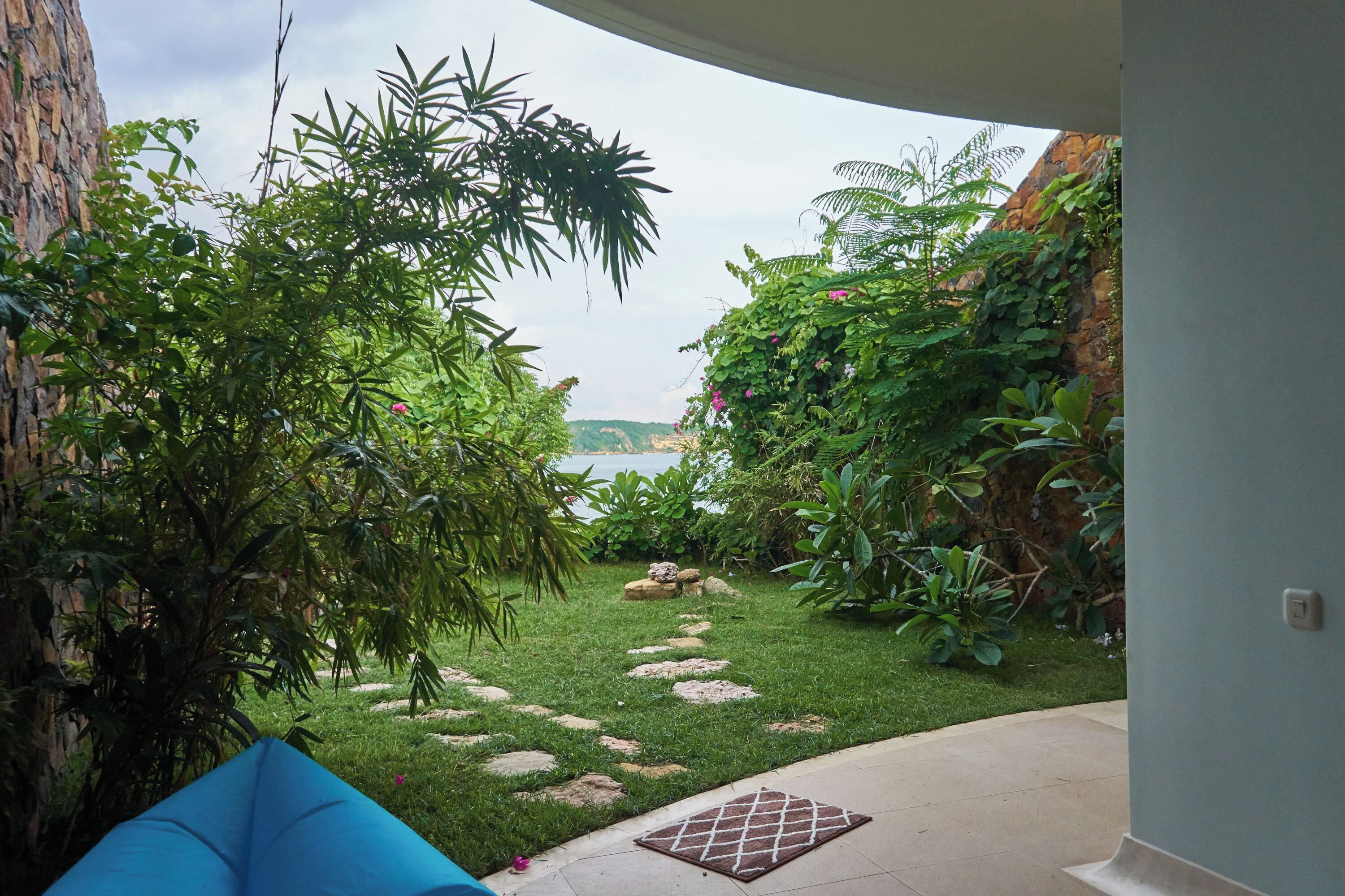 A garden with a stone pathway leading to a body of water, surrounded by lush greenery and a blue chair.