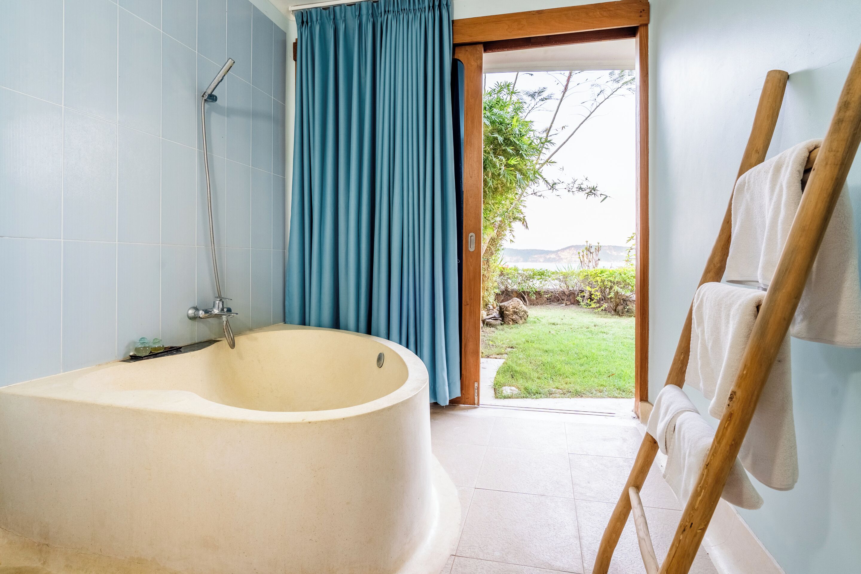 A bathroom with a freestanding bathtub, a wooden ladder with towels, and a view of a garden through an open door.