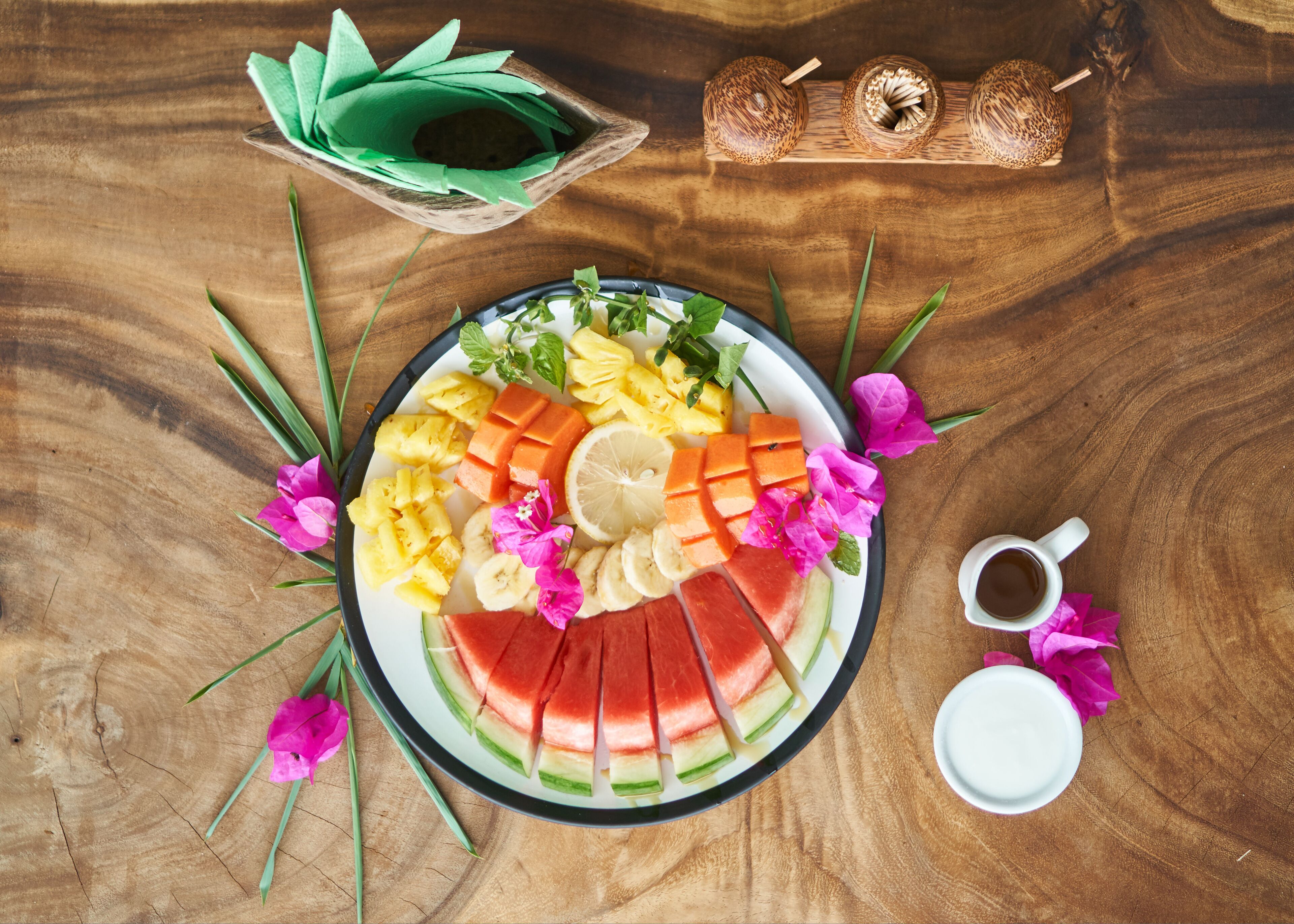 A wooden table with a fruit platter, a cup of coffee, and a small bowl of green leaves.