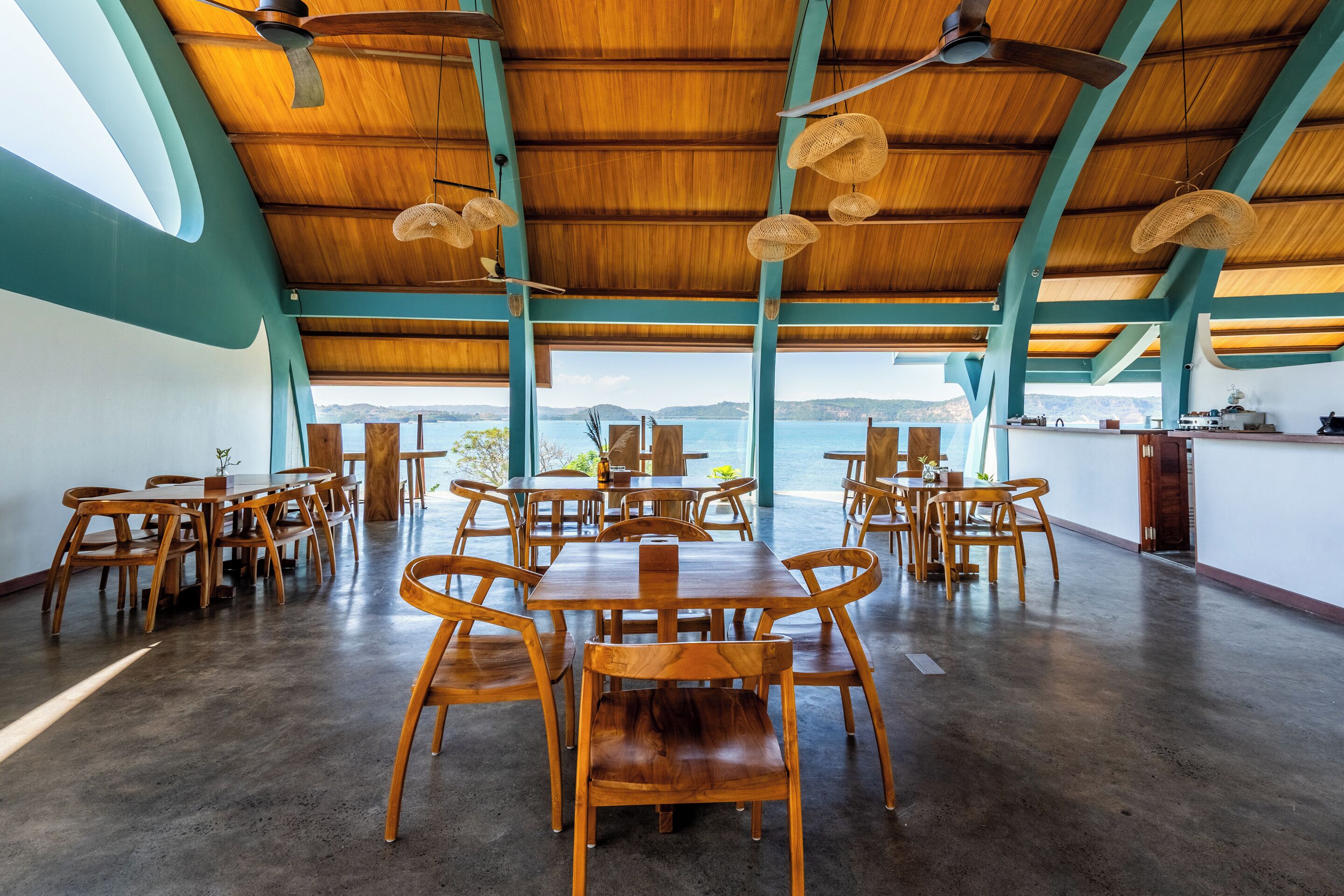 Un comedor amplio con mesas y sillas de madera, un techo alto con lámparas de fibra trenzada y vista al mar.