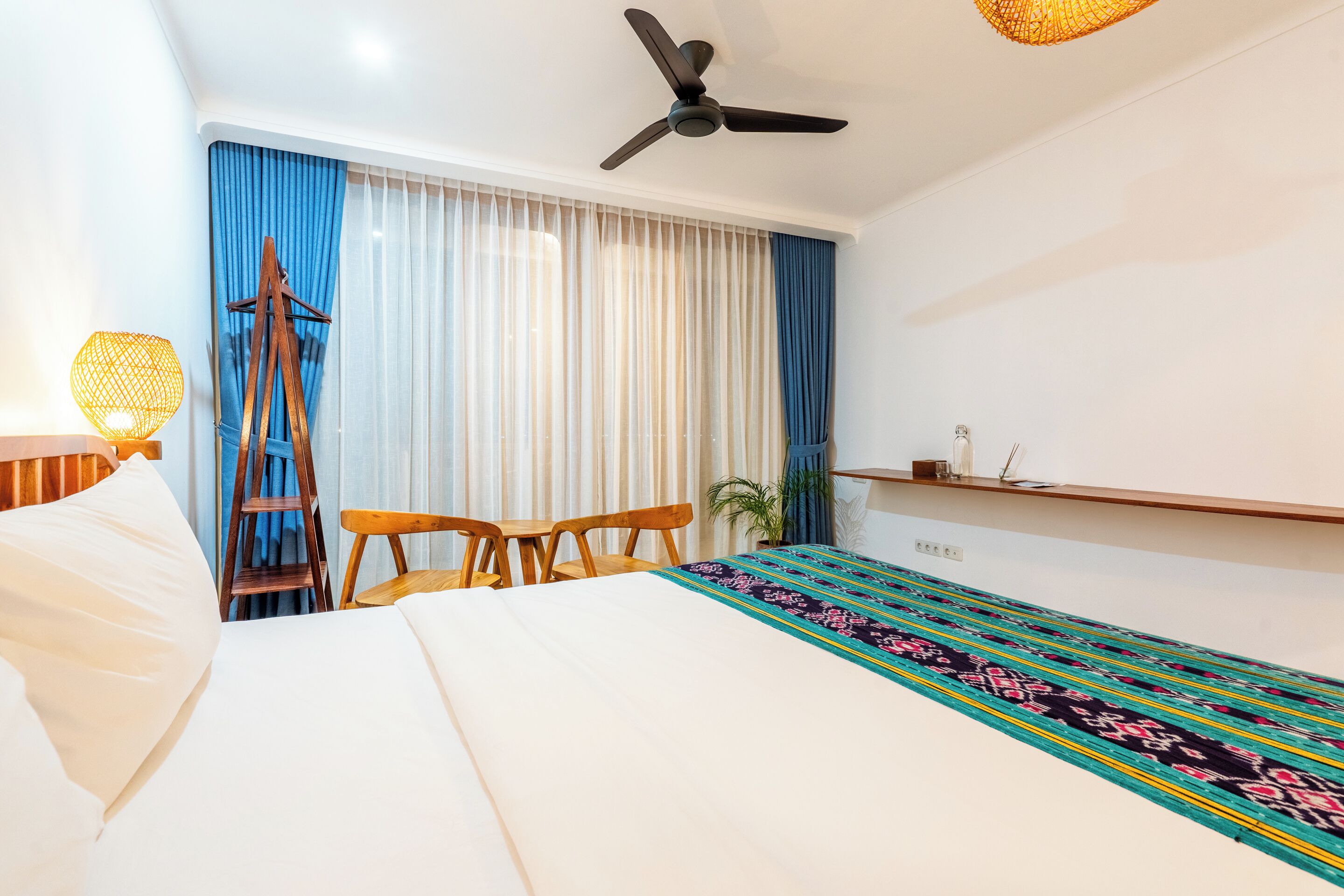 Une chambre d’hôtel moderne équipée d’un lit, d’un bureau, d’une chaise, d’un ventilateur sur pied et d’une fenêtre avec des rideaux.