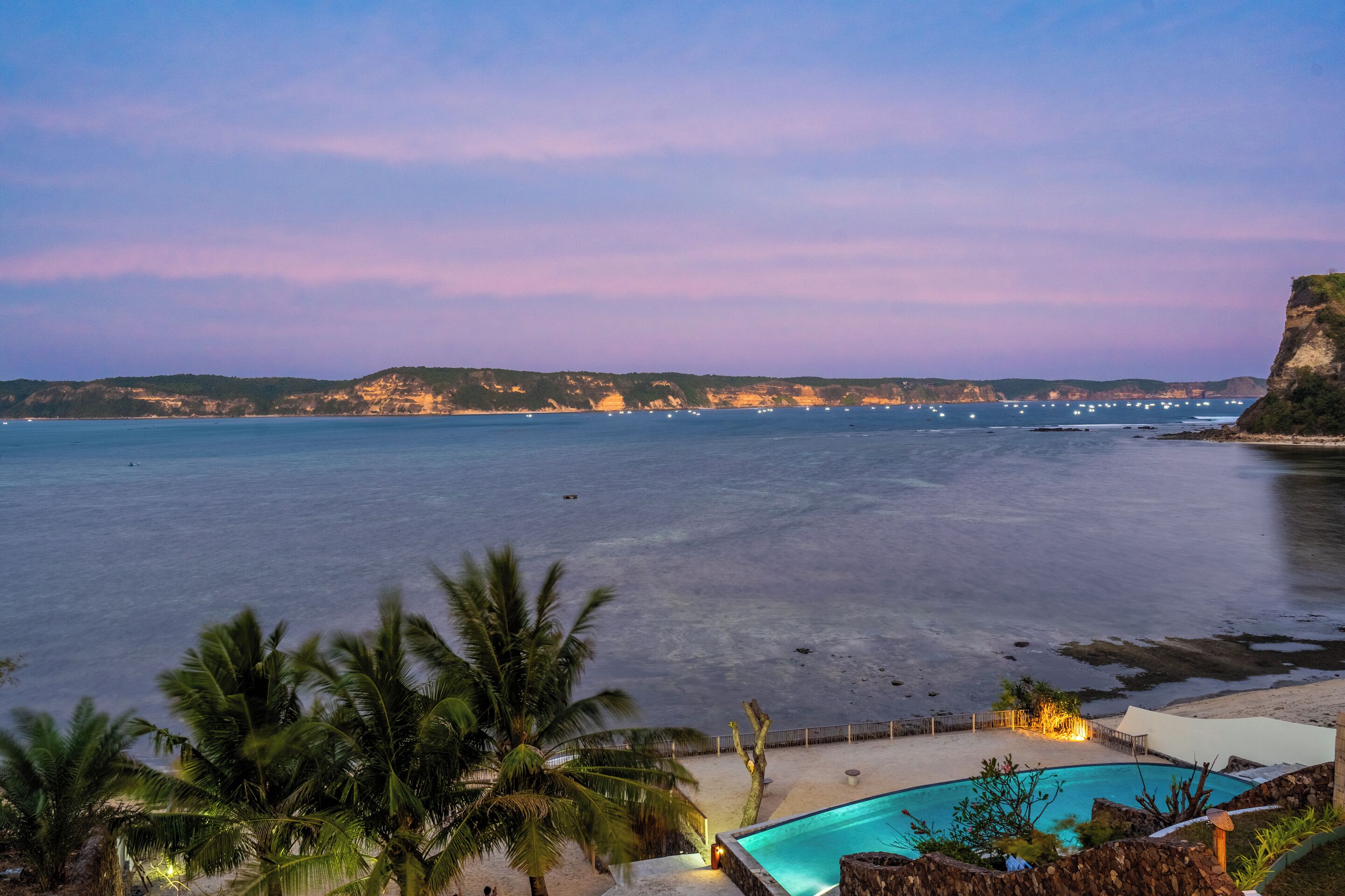 A beachfront property with a swimming pool, palm trees, and a view of the sea and distant cliffs.
