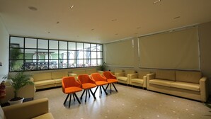Un área de espera con sofás color beige y sillones naranjas, ventanas amplias con vista a mesas al aire libre, y un techo blanco con iluminación empotrada.