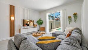 Een moderne woonkamer met een grijze bank, een klein houten bijzettafeltje, een aan een witte muur bevestigde televisie en een glazen schuifdeur die leidt naar een buitenruimte.