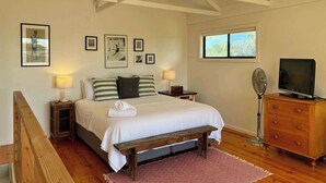 Un dormitorio con cama, mesitas de noche, televisor, ventana y cuadros en la pared.