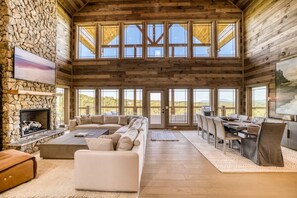 A spacious interior with a stone fireplace, a large sofa, a dining area, and large windows. - Luxury Cabin | Private Pool, Hot Tub & Theater (Smoky Mountains)