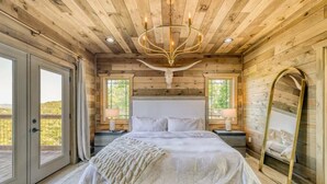 A wooden cabin bedroom with a large bed, a chandelier, and a large window. - Laughing Pines - Indoor pool & theater room (Smoky Mountains)