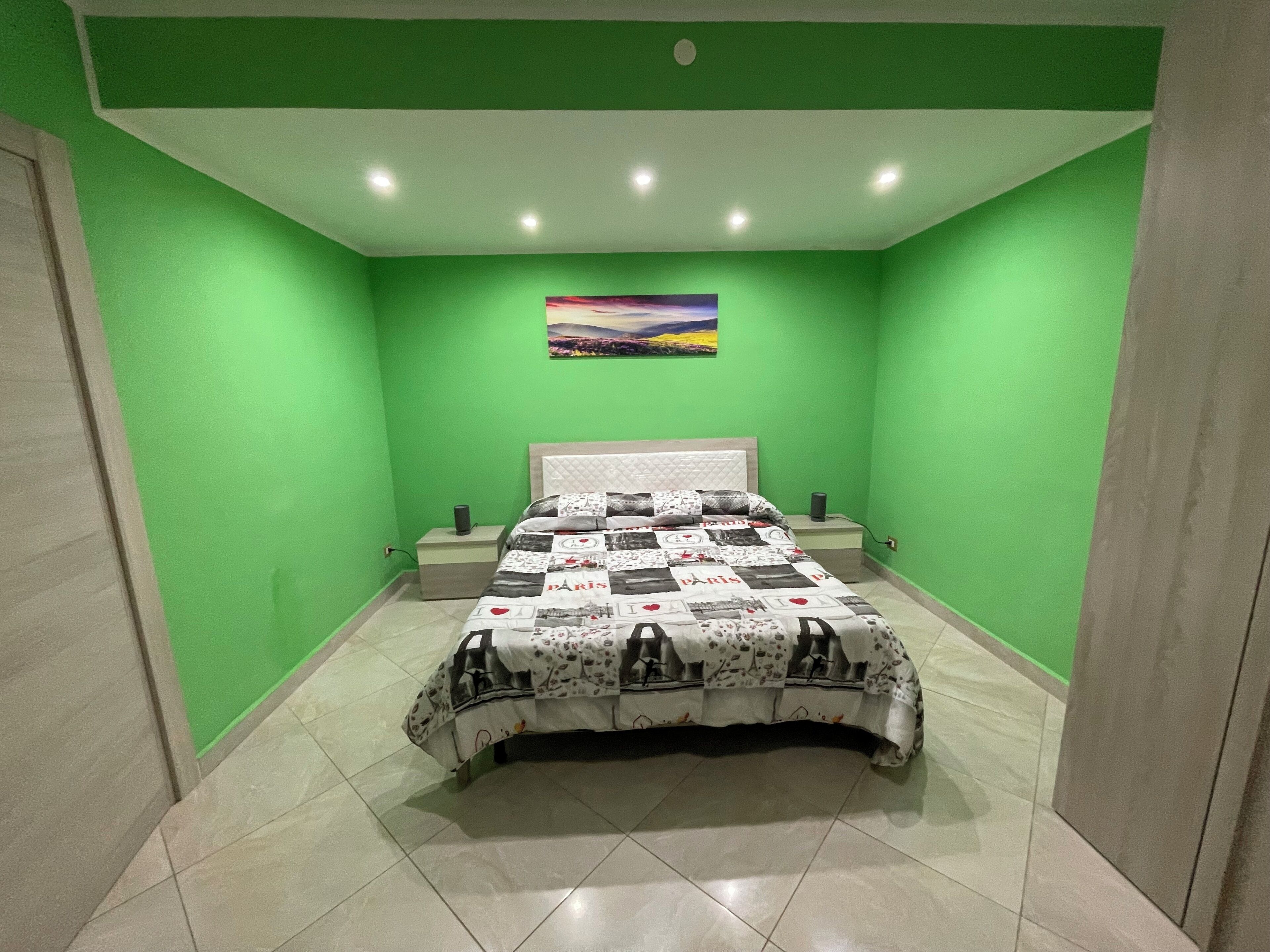 Un dormitorio con paredes verdes, una cama con colcha estampada, una mesita de noche y un cuadro colgado en la pared.