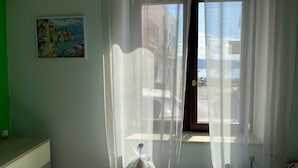 A room with a window covered by sheer curtains, a painting on the wall, and a view of buildings outside. - Cirismiro Torre Faro B&B  (Torre Faro)