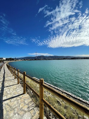A stone pathway with wooden railings beside a calm body of water, leading to a distant mountain range under a clear sky. - Cirismiro Torre Faro B&B  (Torre Faro)