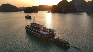 Een grote boot met een vlak dek en meerdere niveaus, afgemeerd in rustig water met een zonsondergang op de achtergrond.