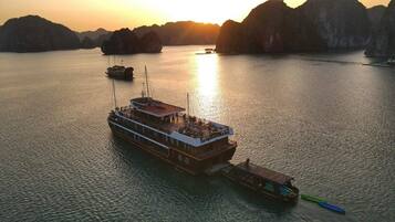 A large boat with a flat deck and multiple levels, docked in calm waters with a sunset in the background.